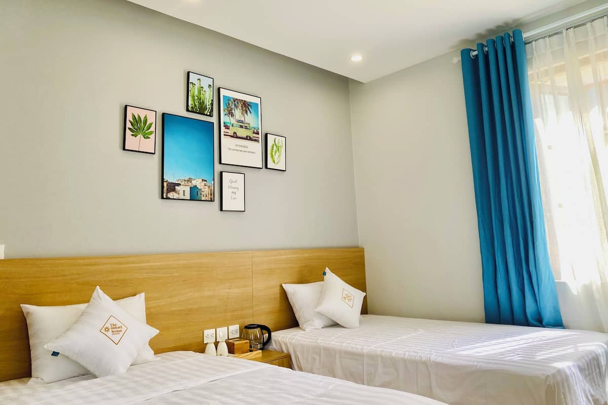 A hotel room with two beds, a wooden headboard, a wall with framed pictures, a window with blue curtains, and a nightstand with a kettle and a box.
