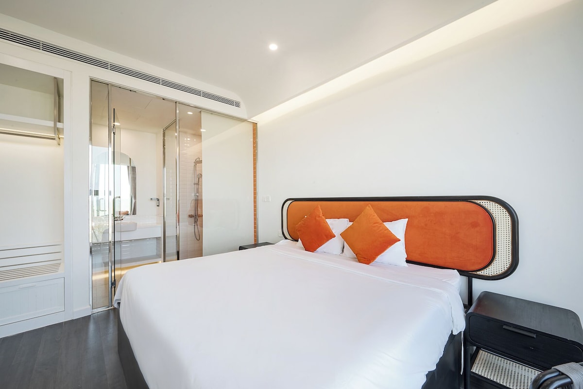 A modern hotel room with a large bed, a glass shower enclosure, and a black nightstand.