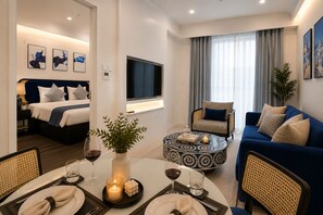A modern hotel room with a bed, a sofa, a dining area with a table set for two, and a TV mounted on the wall.