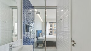 Ein modernes Badezimmer mit einer Duschkabine aus Glas, einem weißen Waschbecken und einem sichtbaren Bett im angrenzenden Zimmer.