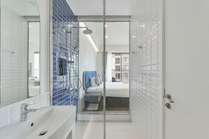 A modern bathroom with a glass shower enclosure, a white sink, and a visible bed in the adjacent room.