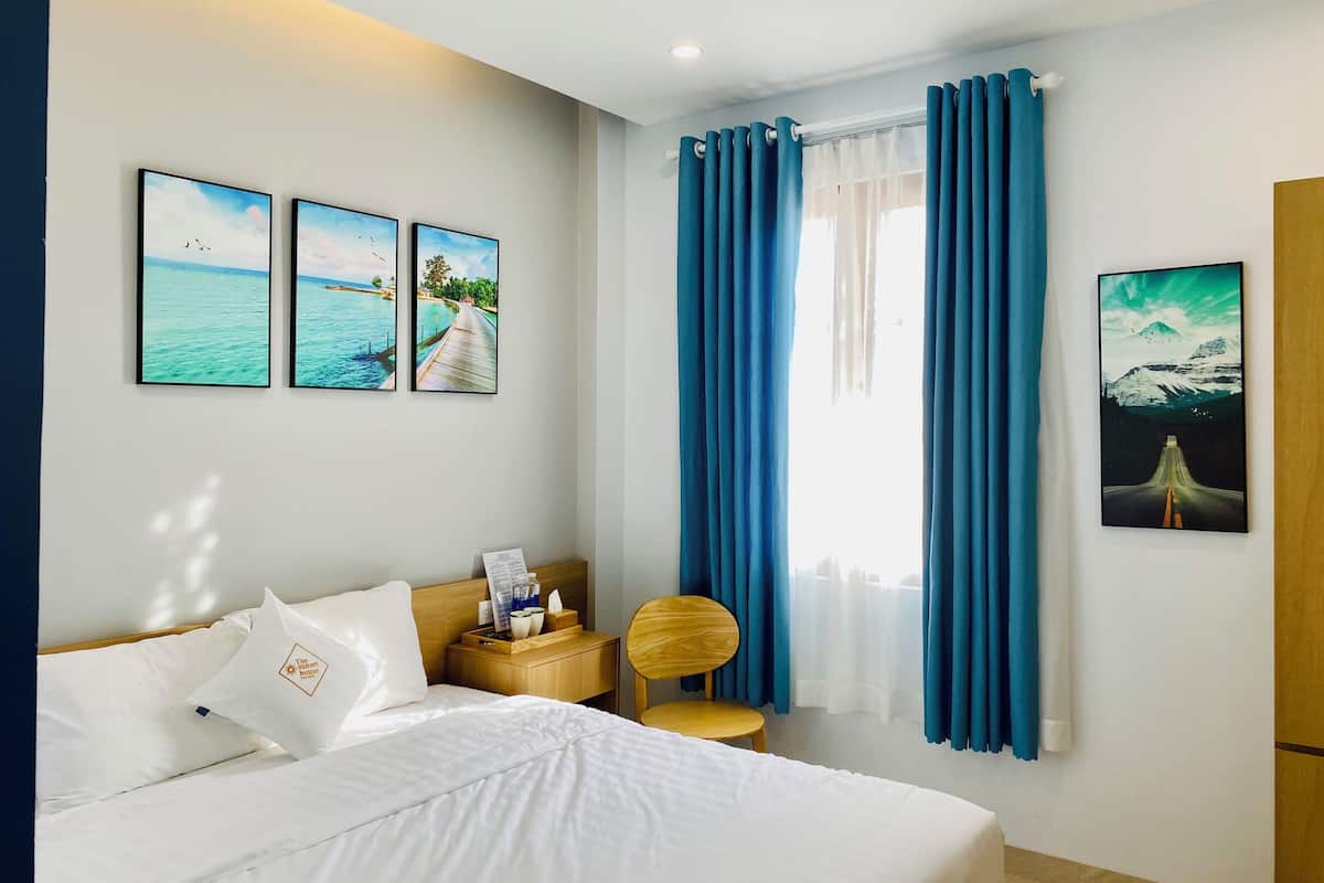 A hotel room with a bed, a wooden nightstand, a chair, and a window with blue curtains. There are three framed pictures on the wall.
