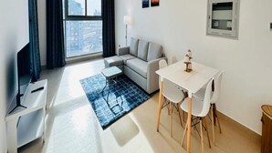 A modern living room with a sofa, a dining table with chairs, a TV, and a large window with curtains. - OYO 1186 Home Orion BLDG Arjan 1 BHK (Dubai)