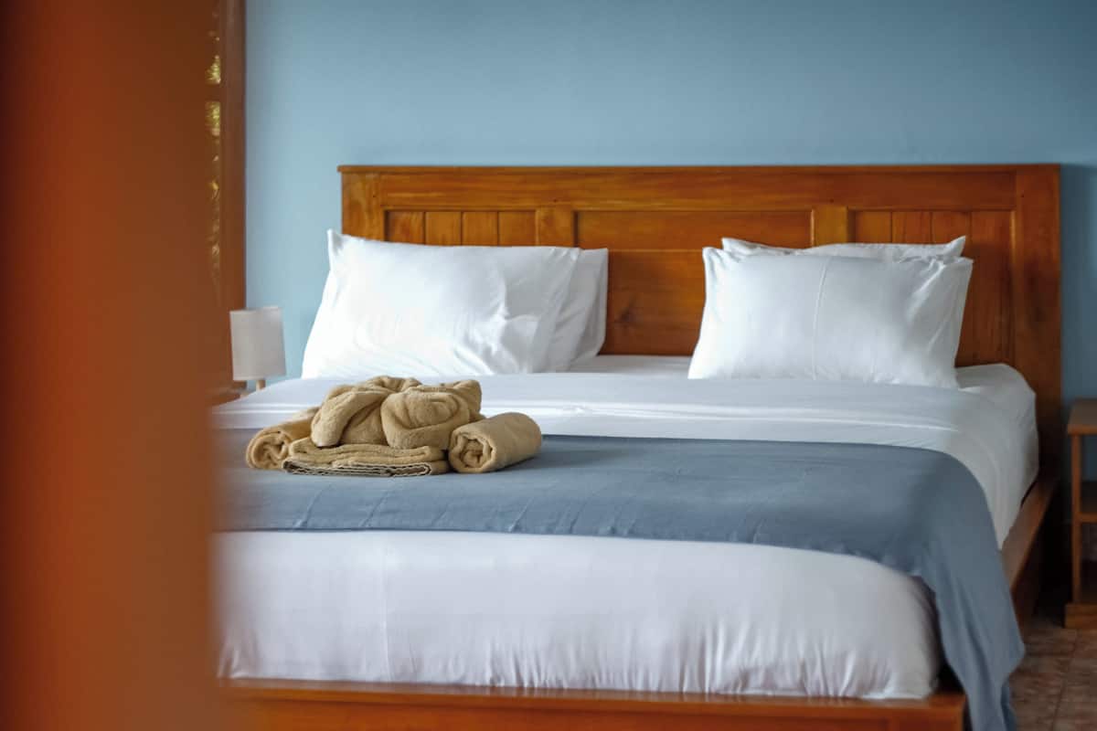 A neatly made bed with white linens and a blue blanket, a wooden headboard, and a rolled-up towel on the bed.