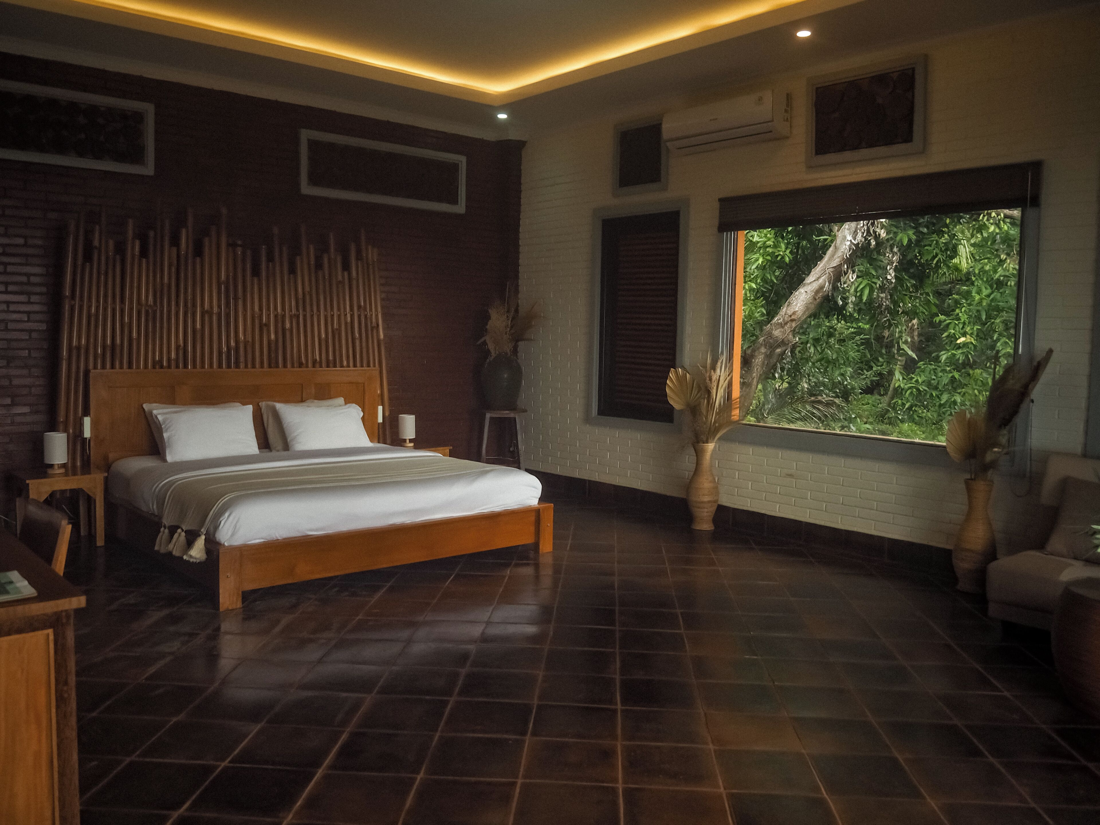 A bedroom with a large bed, a wooden headboard, a window with a view of greenery, and a brick wall.