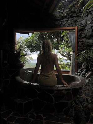 Una persona en una bañera de piedra con vistas a vegetación exuberante y un muro de piedra.