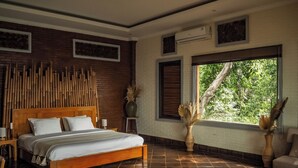 A spacious bedroom with a large bed, a wooden headboard, a brick wall, and a window with a view of greenery. - Kailash Lodge  (Tejakula)