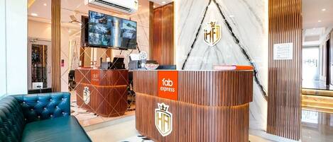 A hotel reception area with a modern design, featuring a reception desk, a large flat-screen TV, and a comfortable seating area with a blue sofa.