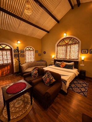 A cozy bedroom with a large bed, a sofa, and a small table. There are two windows with curtains, a chandelier, and wall-mounted lamps. - La Address Retreat (Ootacamund)