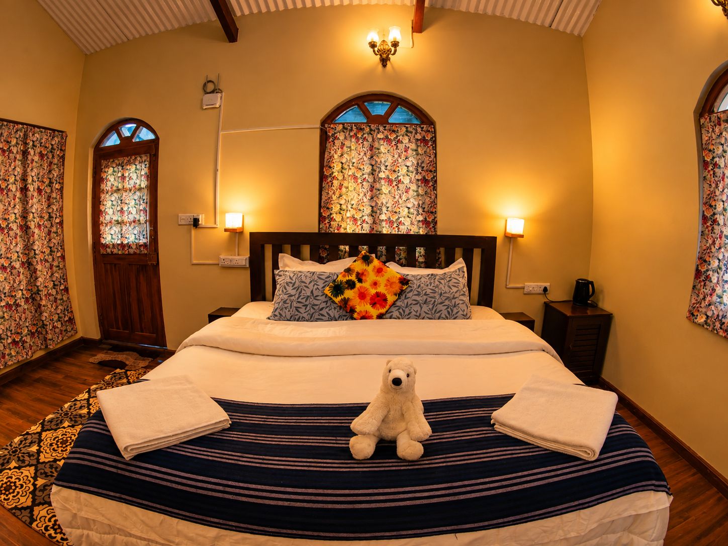 A hotel room with a bed, a teddy bear, a window with floral curtains, and a chandelier.