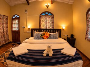 A hotel room with a bed, a teddy bear, a window with floral curtains, and a chandelier. - La Address Retreat (Ootacamund)