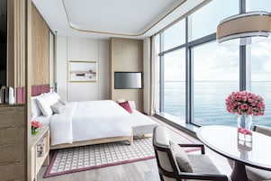 A modern hotel room with a large bed, a TV, a dining table with chairs, and a view of the sea.