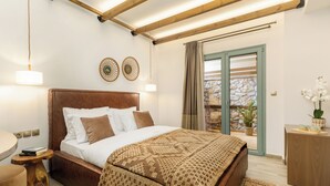 Une chambre à coucher avec une tête de lit en bois, un grand lit recouvert d’une literie à motifs, une table de chevet avec des livres, et une vue sur l’extérieur grâce à une porte vitrée.