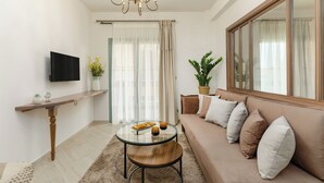 Un salon comprenant un canapé, une table basse, un téléviseur fixé au mur, un lustre et une fenêtre avec des rideaux.