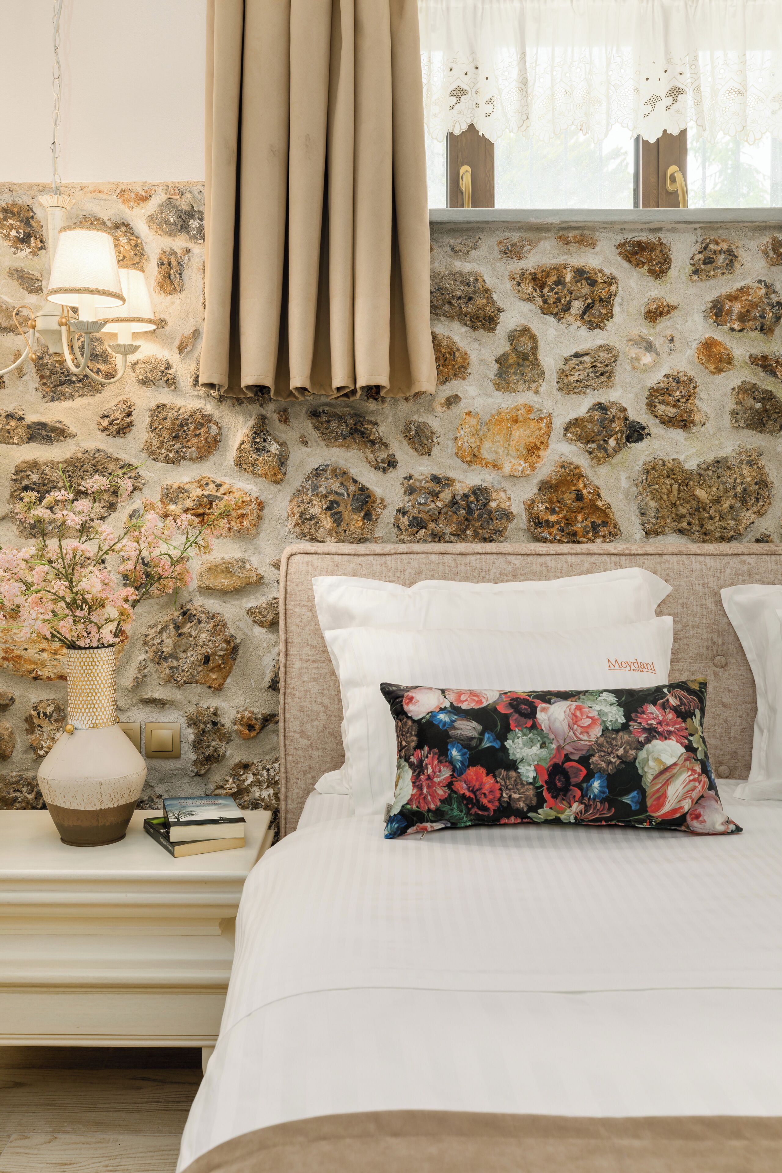 Un dormitorio con pared de piedra, una cama con ropa de cama blanca, un cojín floral, una mesita de noche con una lámpara y un jarrón con flores.