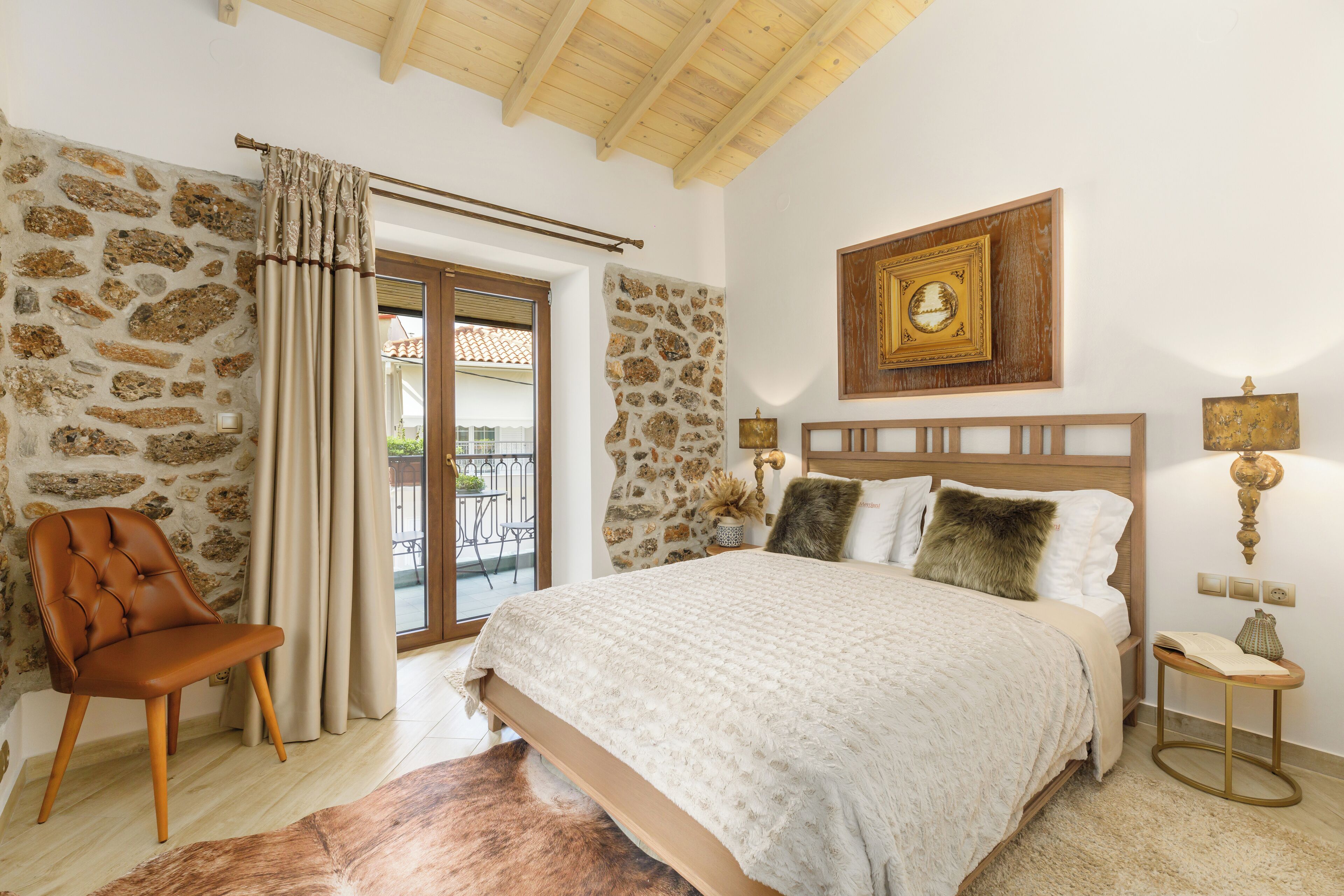 Un dormitorio con pared de piedra, techo de madera, cama con cubrecama blanco, una silla, una mesita de noche con lámpara y un cuadro enmarcado en la pared.