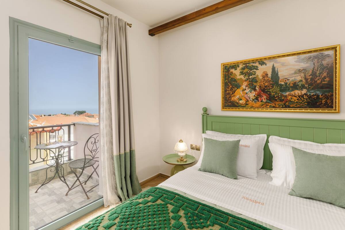 A bedroom with a green headboard, a painting on the wall, a balcony with a table and chairs, and a view of the sea.