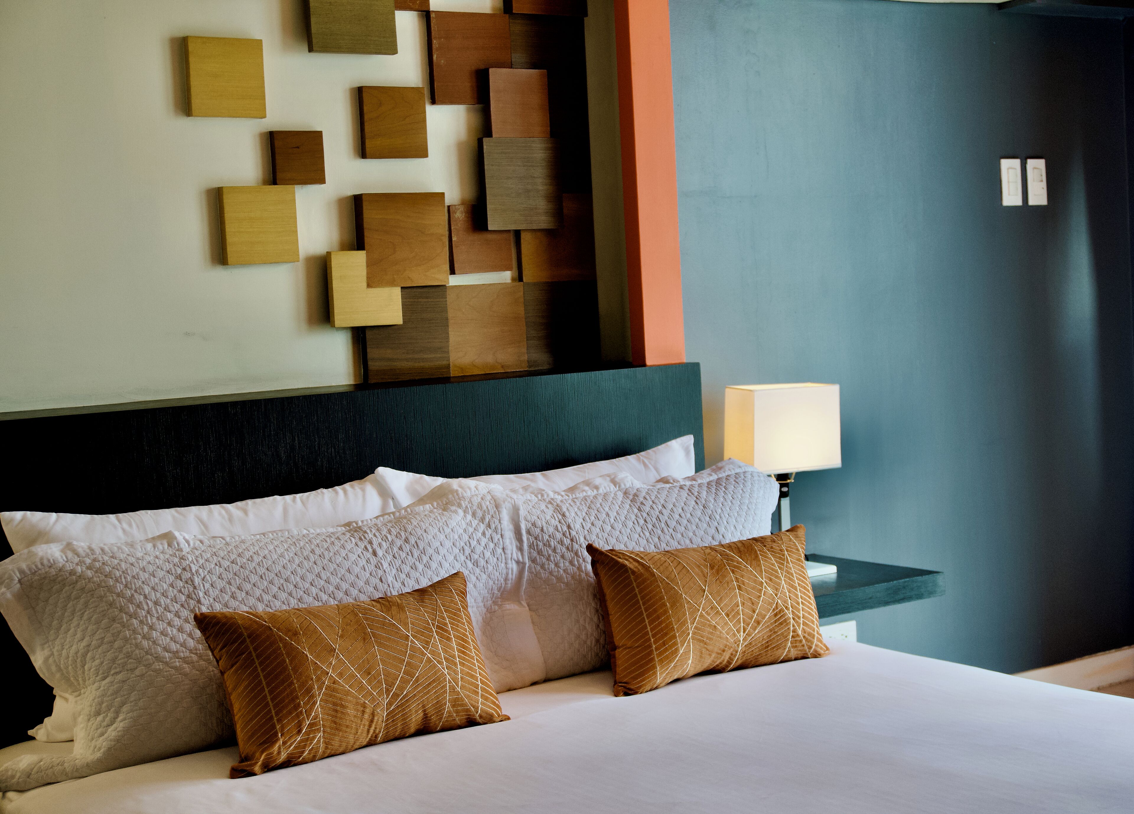 Een netjes opgemaakt bed met witte en bruine kussens, een donker hoofdbord en een muur met een geometrisch houten paneelpatroon.
