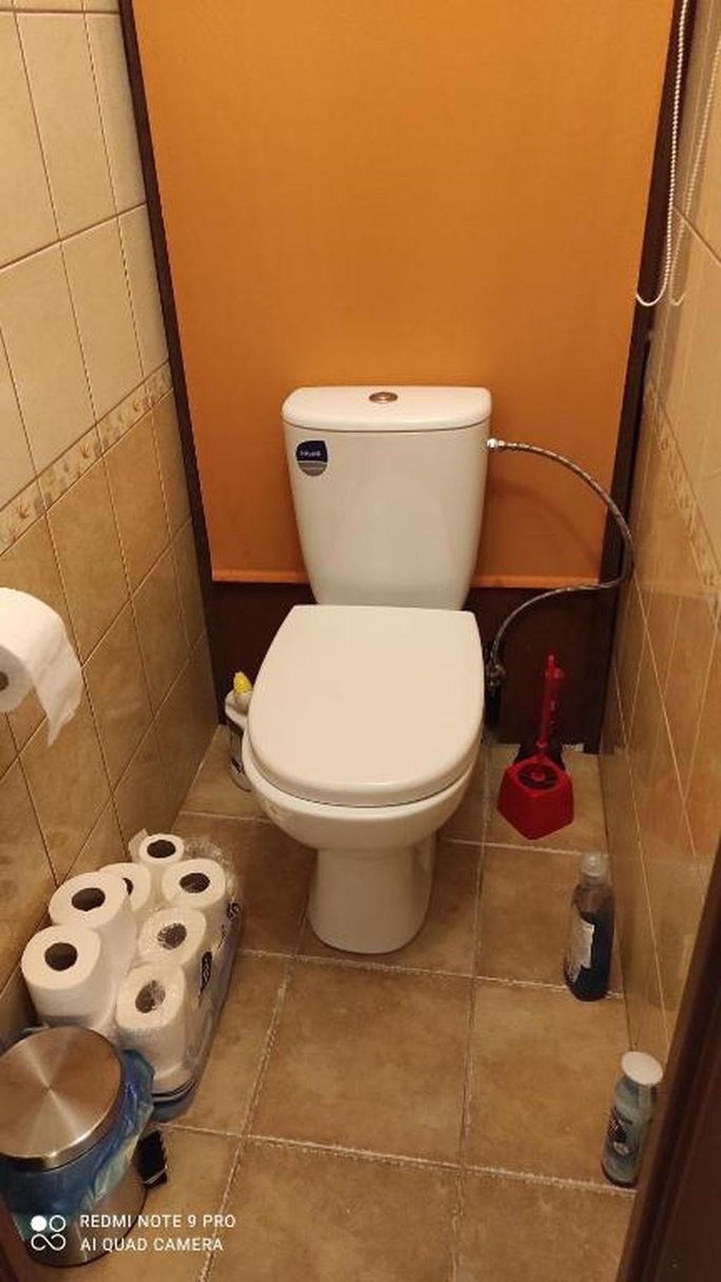 Une petite salle de bain avec des toilettes, un rouleau de papier toilette, une serpillière rouge et une bouteille de produit nettoyant.