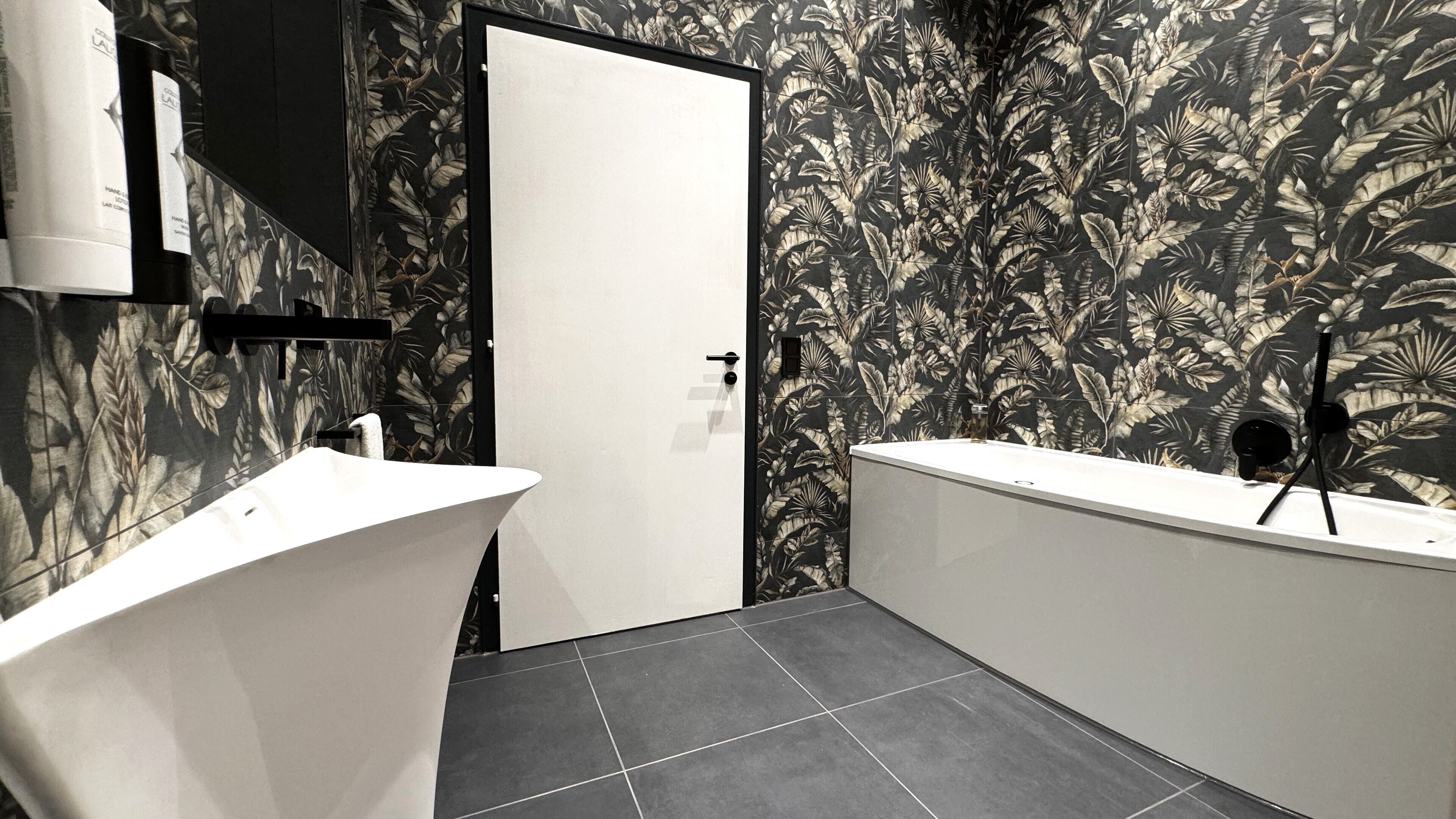 Ein modernes Badezimmer mit einer weißen Badewanne, einer separaten Duschkabine und einem WC. Die Wände sind mit einem tropischen Blattmuster verziert.