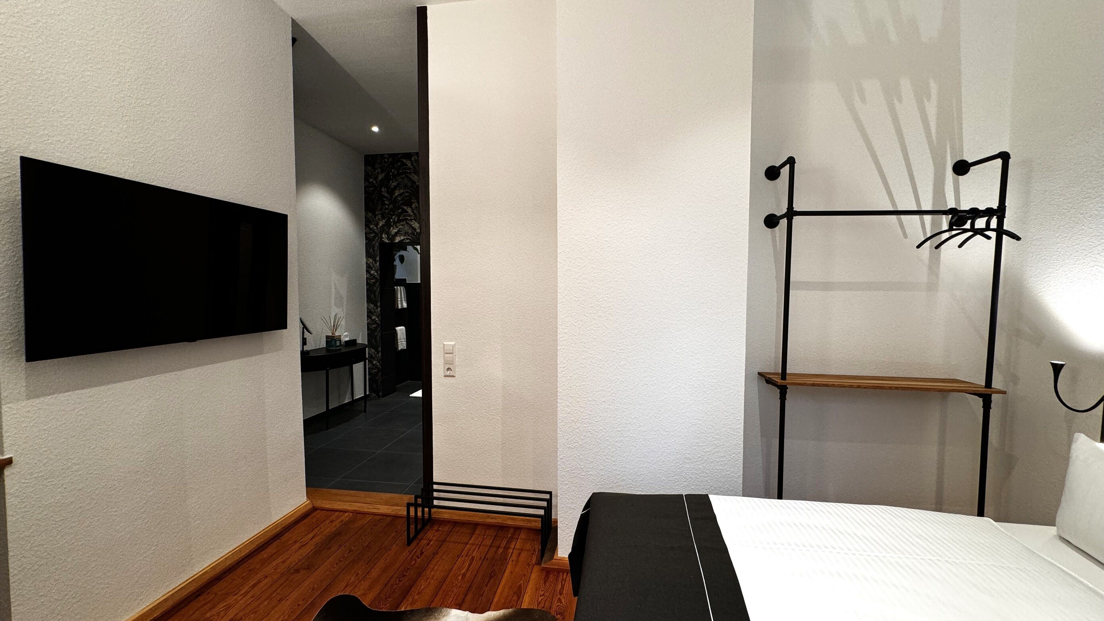 Una habitación de hotel moderna con una televisión de pantalla plana montada en la pared, suelo de madera, un banco negro, una cama con colchón blanco y un perchero metálico con ganchos.
