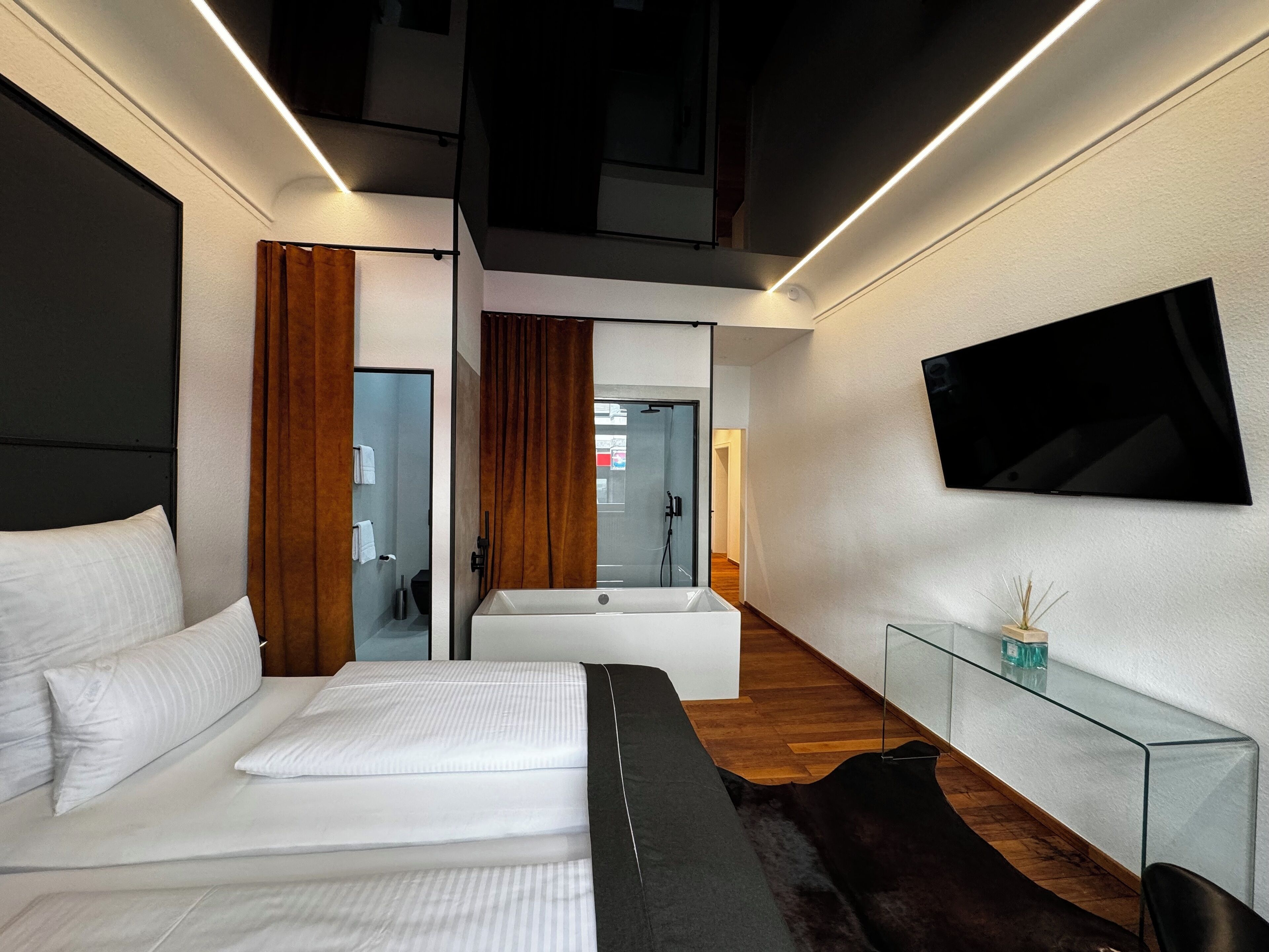 Une chambre d’hôtel moderne dotée d’un grand lit, d’une table de chevet en verre, d’un téléviseur fixé au mur et d’une salle de bain avec baignoire et douche.