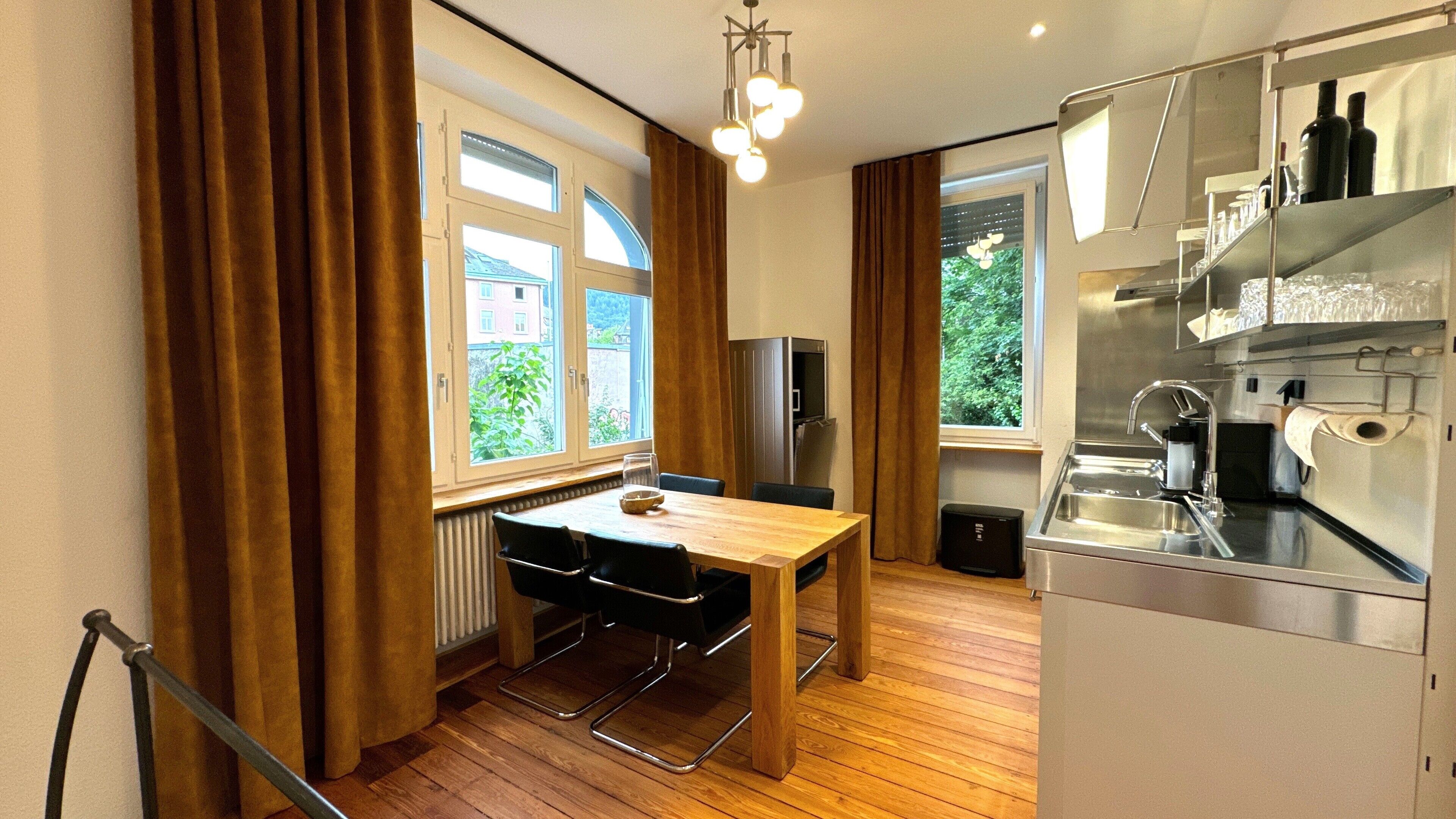 A compact kitchen with a dining area, wooden flooring, and large windows with curtains.