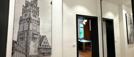 Un pasillo con dibujos arquitectónicos enmarcados en las paredes, suelo de madera y una puerta que da a una habitación con una mesa y sillas.