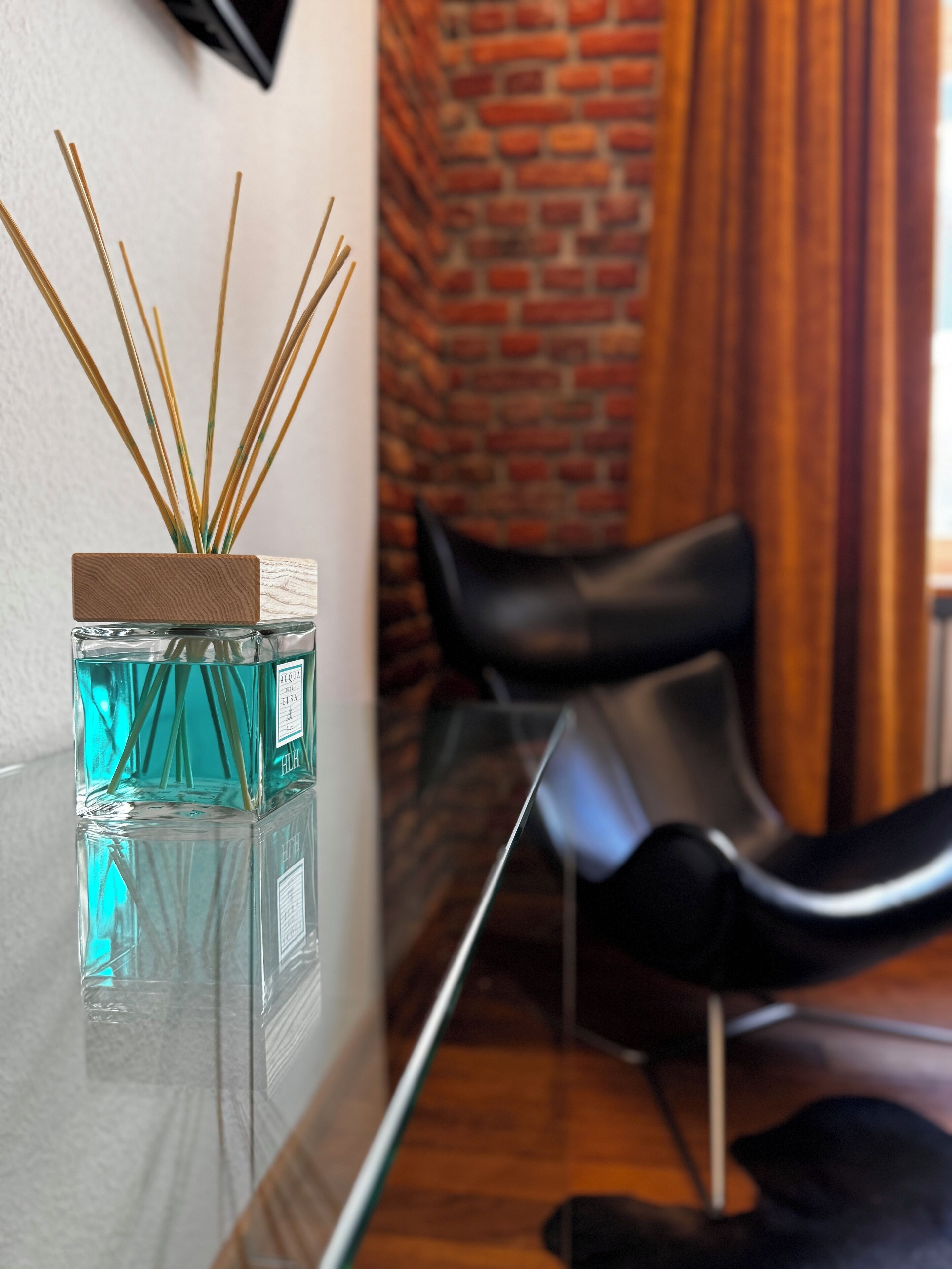 Een glazen tafel met een flesje diffuser en een houten blok. Een zwarte stoel is op de achtergrond te zien.