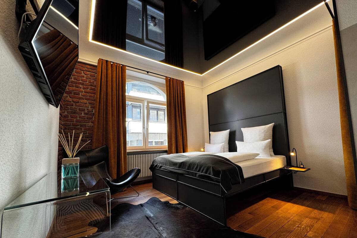 A modern hotel room with a black leather bed, a glass desk, a leather chair, a black cowhide rug, a brick wall, and a window with curtains.