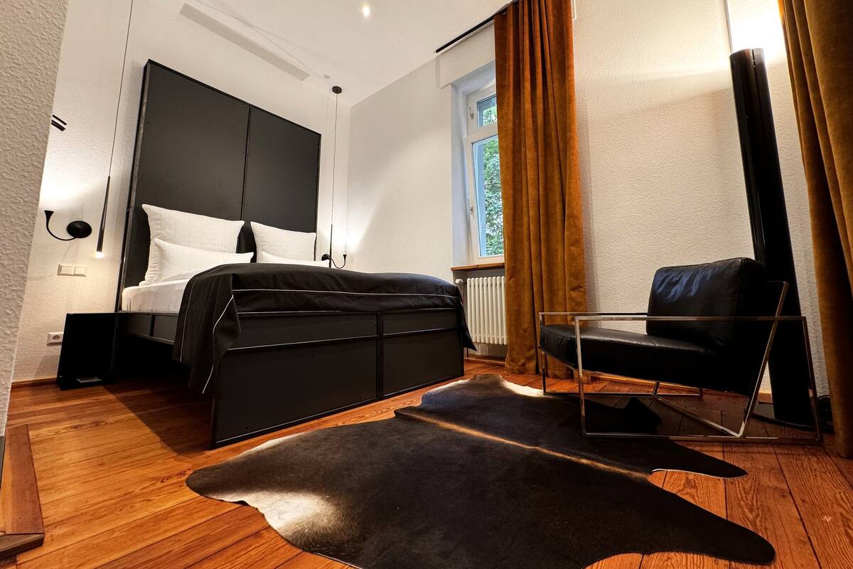 A modern bedroom with a black bed, a wooden floor, a black leather chair, and a large window with curtains.