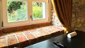 Una habitación con ventana a zonas verdes, una mesa con un libro y un control remoto, y una pared de piedra.