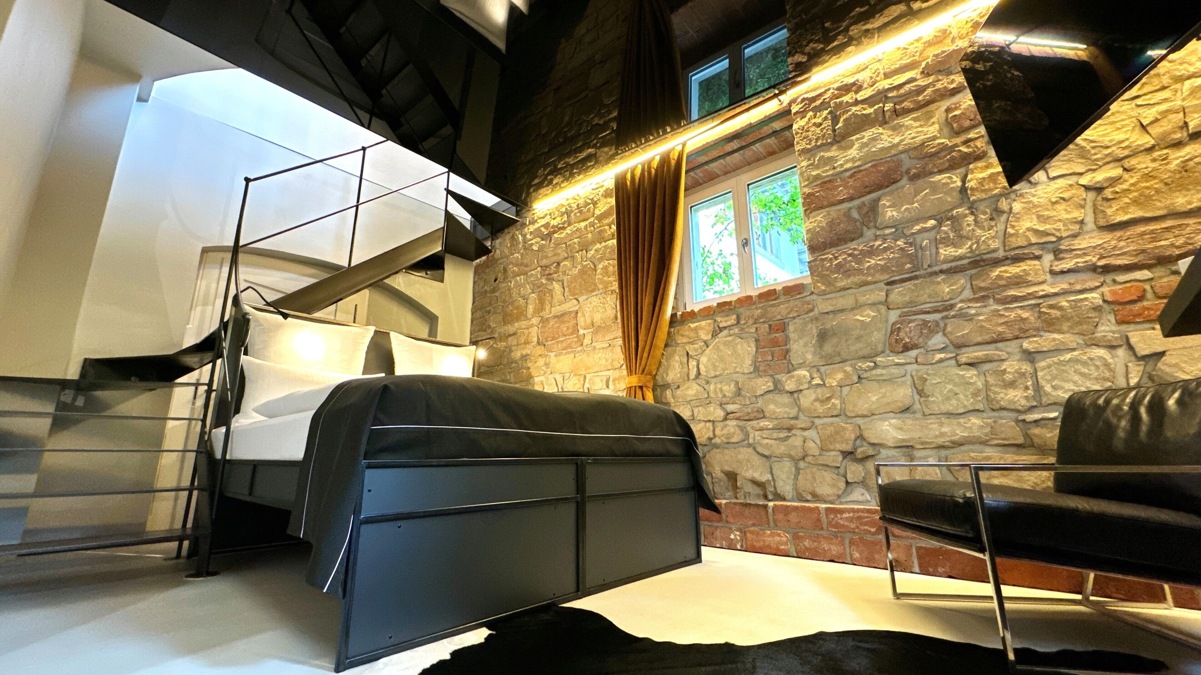 A modern room with a stone wall, a bed with a black canopy, a leather sofa, and a staircase.