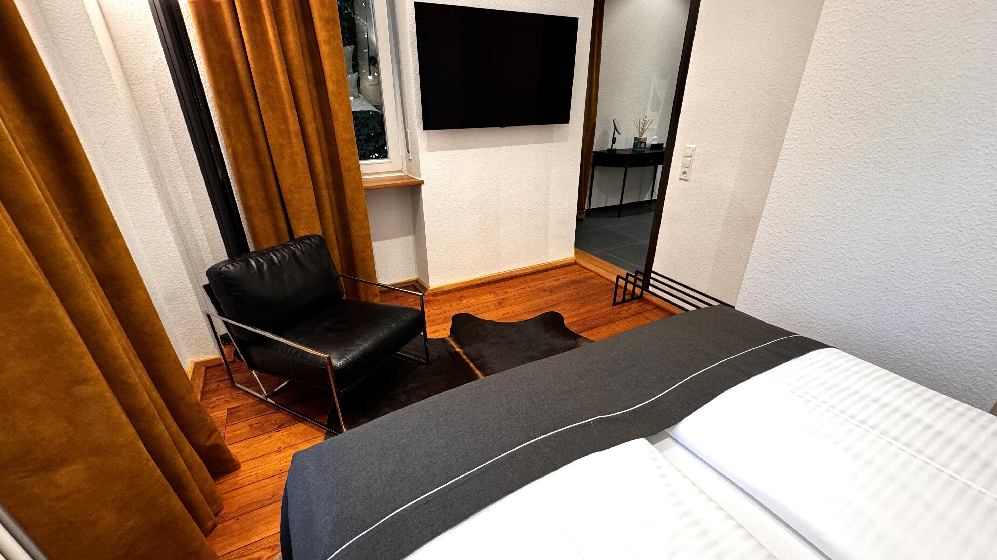 Habitación de hotel con cama, un sillón de cuero negro, una alfombra pequeña y un televisor de pantalla plana montado en la pared.