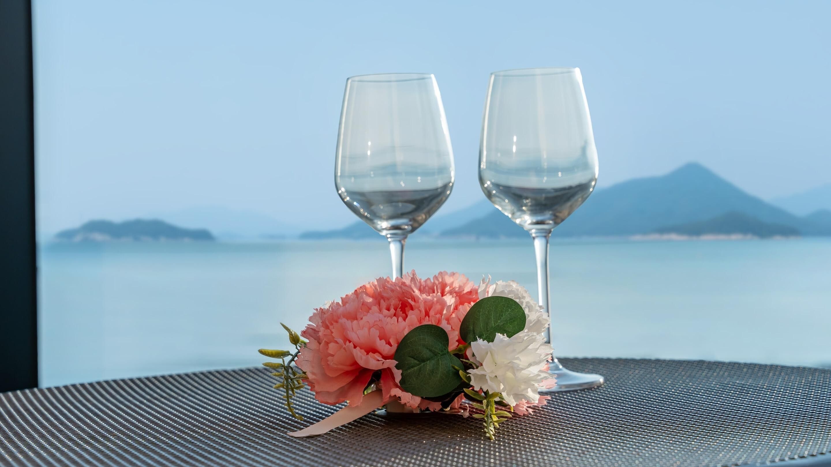 Zwei Weingläser und eine Blumenkomposition auf einem Tisch mit Blick auf das Meer und die Berge.