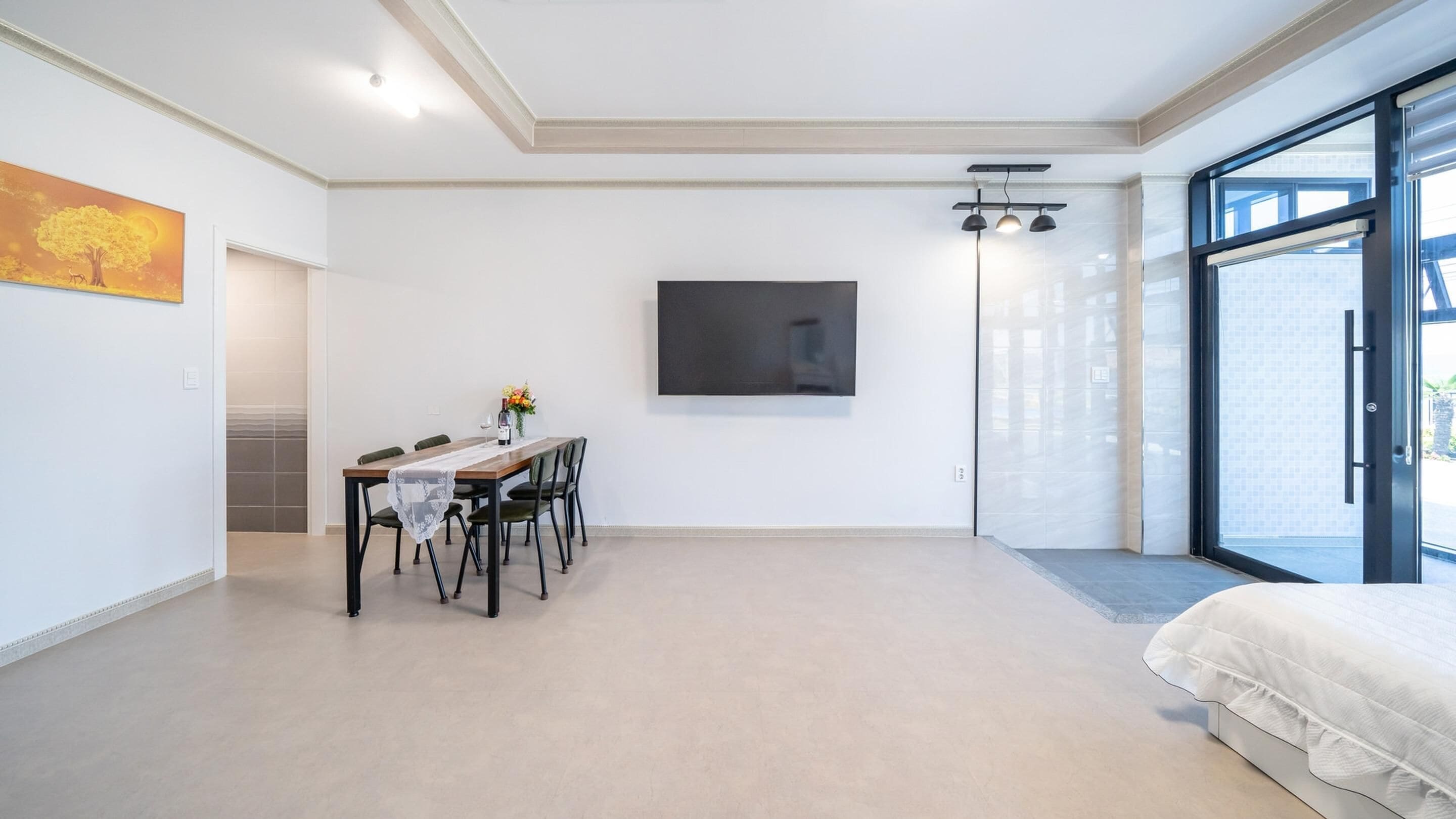 A modern bedroom with a large bed, a dining area with a table and chairs, a flat-screen TV mounted on the wall, and a sliding glass door leading to an outdoor area.