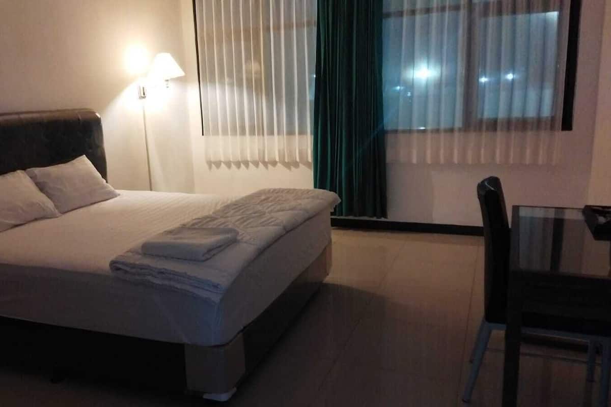 A neatly made bed with pillows and a folded blanket, a bedside lamp, a chair, and a window with blinds.
