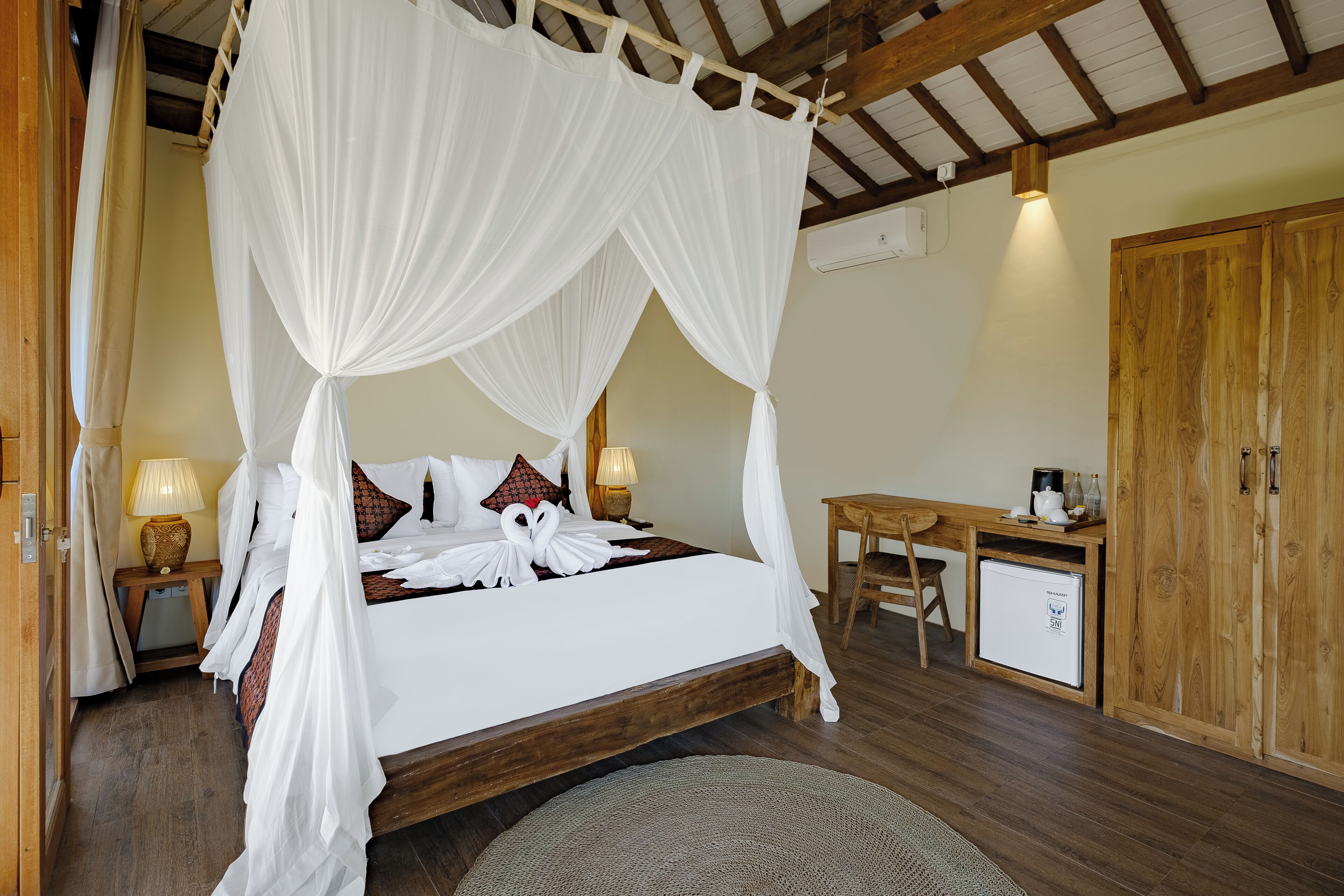 A four-poster bed with white linens, a wooden desk, a small refrigerator, and a wooden wardrobe.