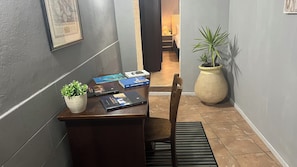 Un escritorio con libros, una planta en maceta y una silla en un pasillo con suelo de baldosas y una planta grande en maceta.