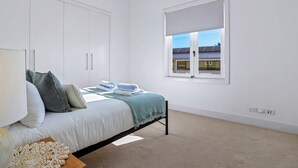 A bedroom with a bed, pillows, a bedside table, a window with blinds, and a view of a building outside. - Elegance On The River, Noosa Waters (Noosaville)