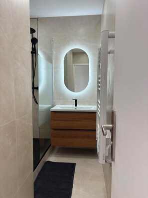 Un bagno moderno con cabina doccia in vetro, un mobile lavabo in legno, uno specchio rotondo con illuminazione integrata e un tappeto nero sul pavimento.