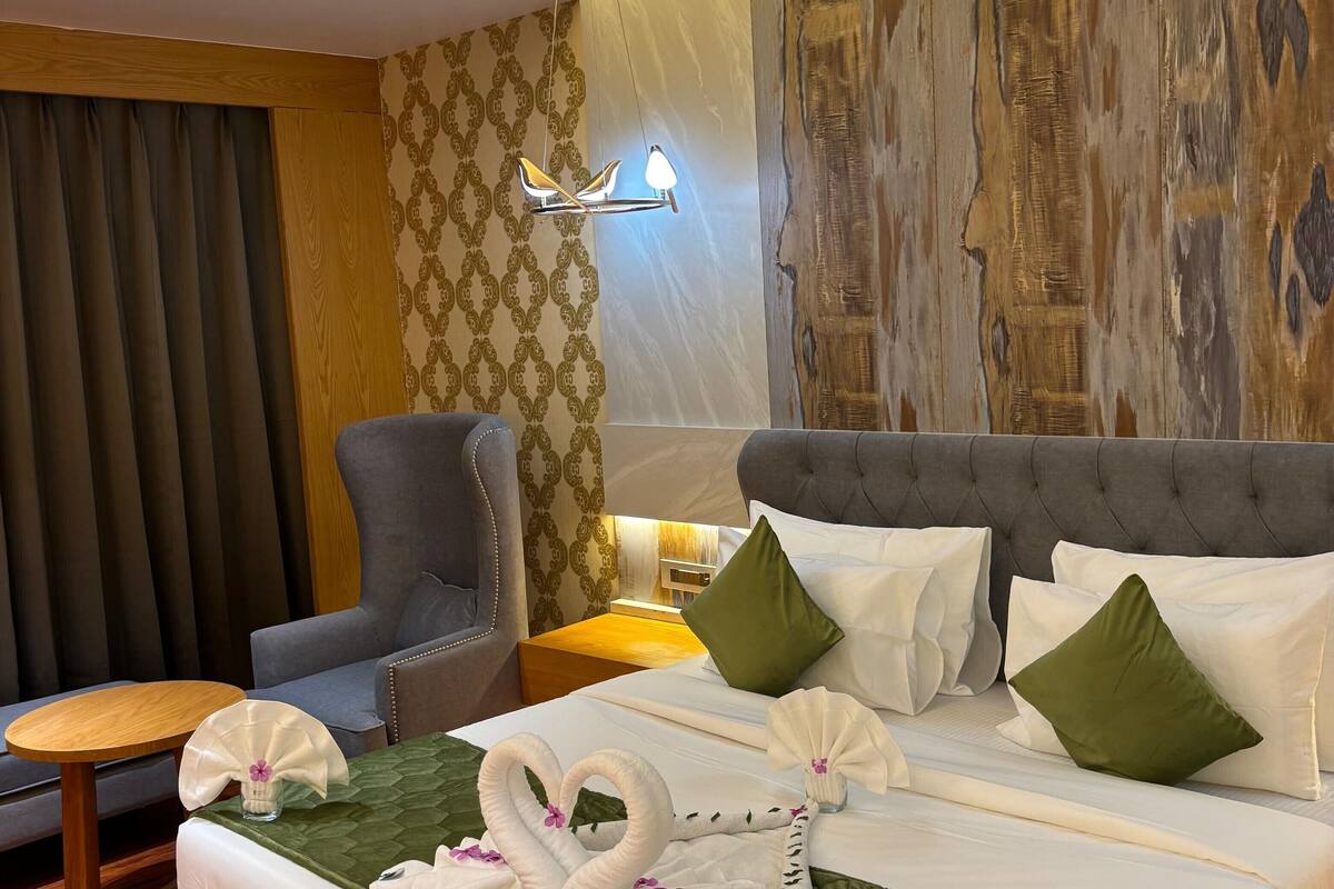 A hotel room with a neatly made bed, a wooden nightstand, a grey armchair, and a small wooden table.