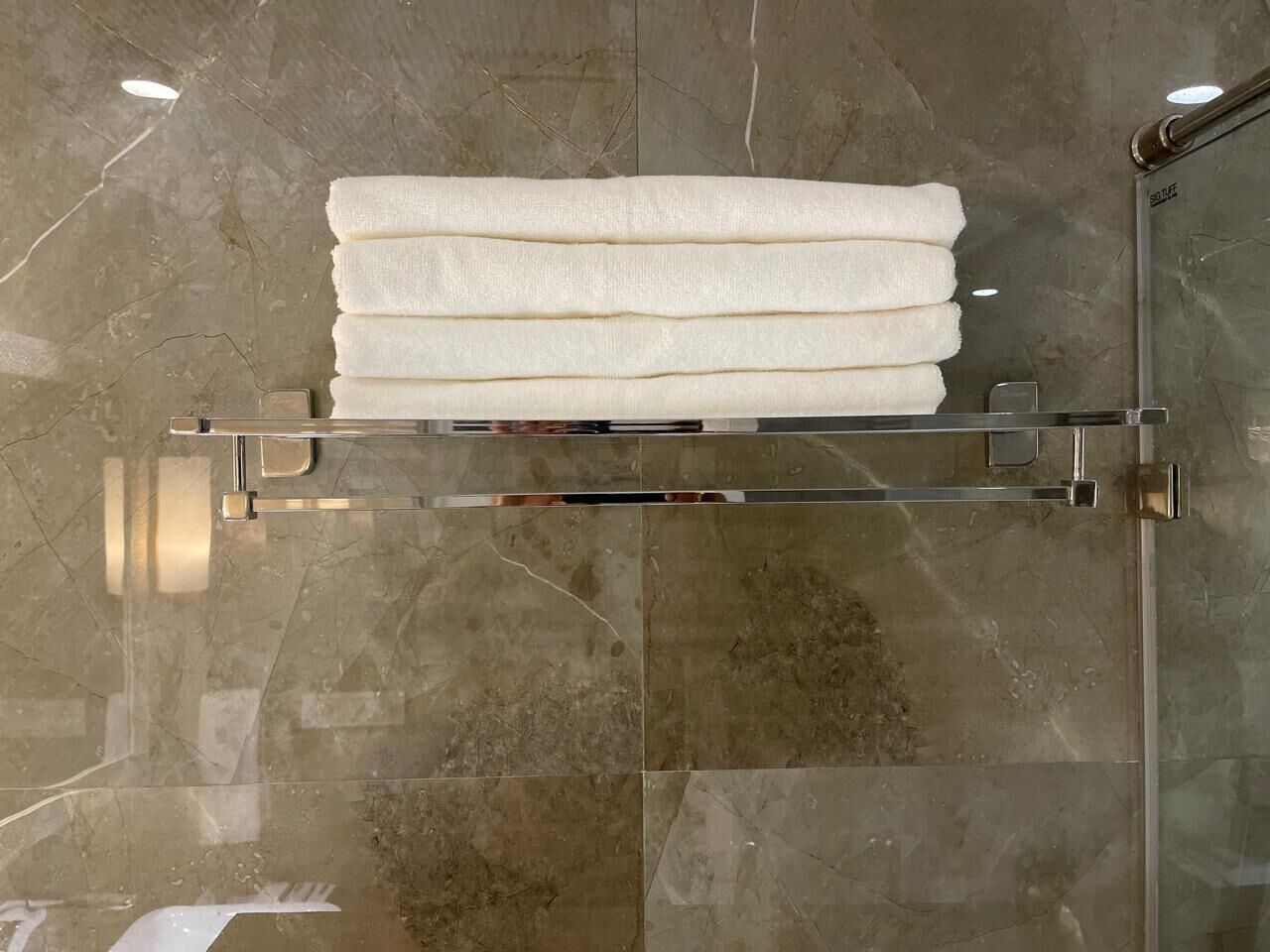 Un bany amb un envà de dutxa de vidre, un suport per tovalloles blanc amb tovalloles doblegades i un paviment de marbre.