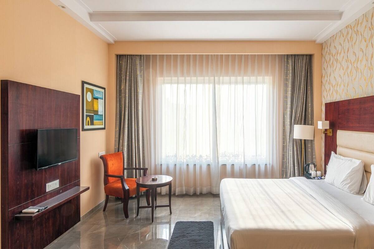 A hotel room with a bed, a TV mounted on the wall, a chair, a small table, and a window with curtains.
