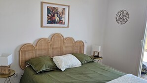 A bedroom with a bed, a wicker headboard, a framed artwork, a bedside table, and a floor lamp. - Appartement Neuf de 84 m2 Avec Terrasse de 60 m2 vue mer (L'Île-Rousse)