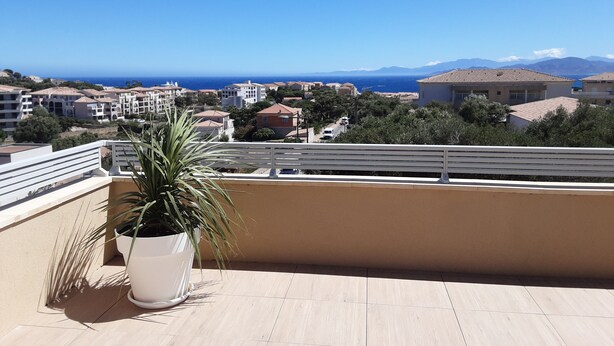 A balcony with a potted plant, overlooking a coastal town with buildings and the sea. - Appartement Neuf de 84 m2 Avec Terrasse de 60 m2 vue mer (L'Île-Rousse)