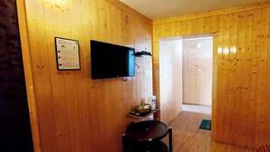 A wooden-paneled room with a flat-screen TV mounted on the wall, a small table with a bowl of fruit, and a door leading to another room. - Ikraam Inn (Srinagar)
