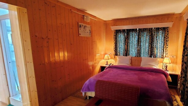 A wooden-paneled bedroom with a bed, two bedside lamps, and a window with patterned curtains. - Ikraam Inn (Srinagar)
