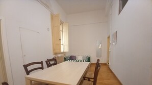 Uma sala de jantar com uma mesa comprida e cadeiras, um radiador, uma janela com portaria aberta e um espelho na parede.