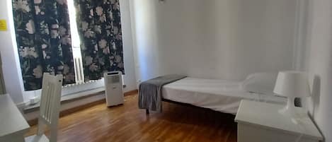 A small, sparsely furnished room with a bed, a chair, a desk, and a radiator.